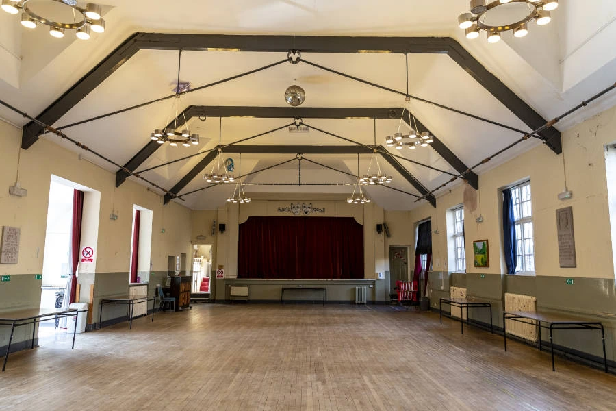 Moor Pool’s empty Main Hall, with a wooden floor, exposed roof beams and a red-curtained stage