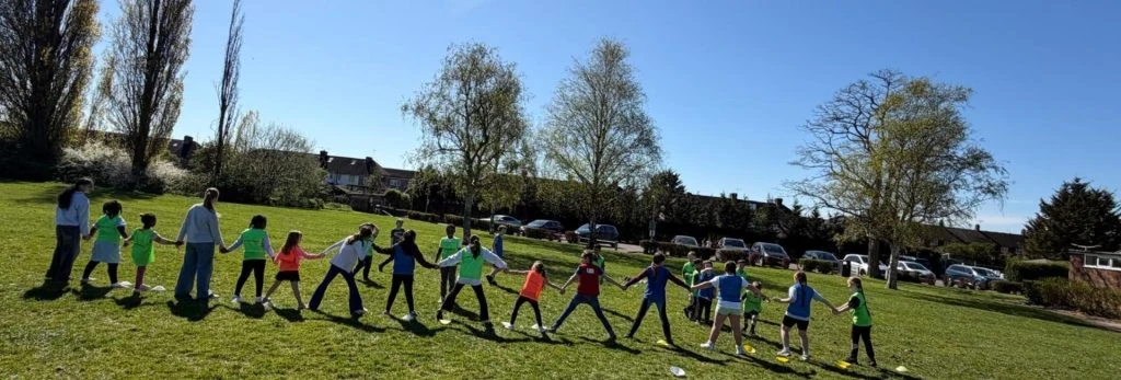 Children and adults holding hands in a long line on grass during an outdoor Stanwell Events activity