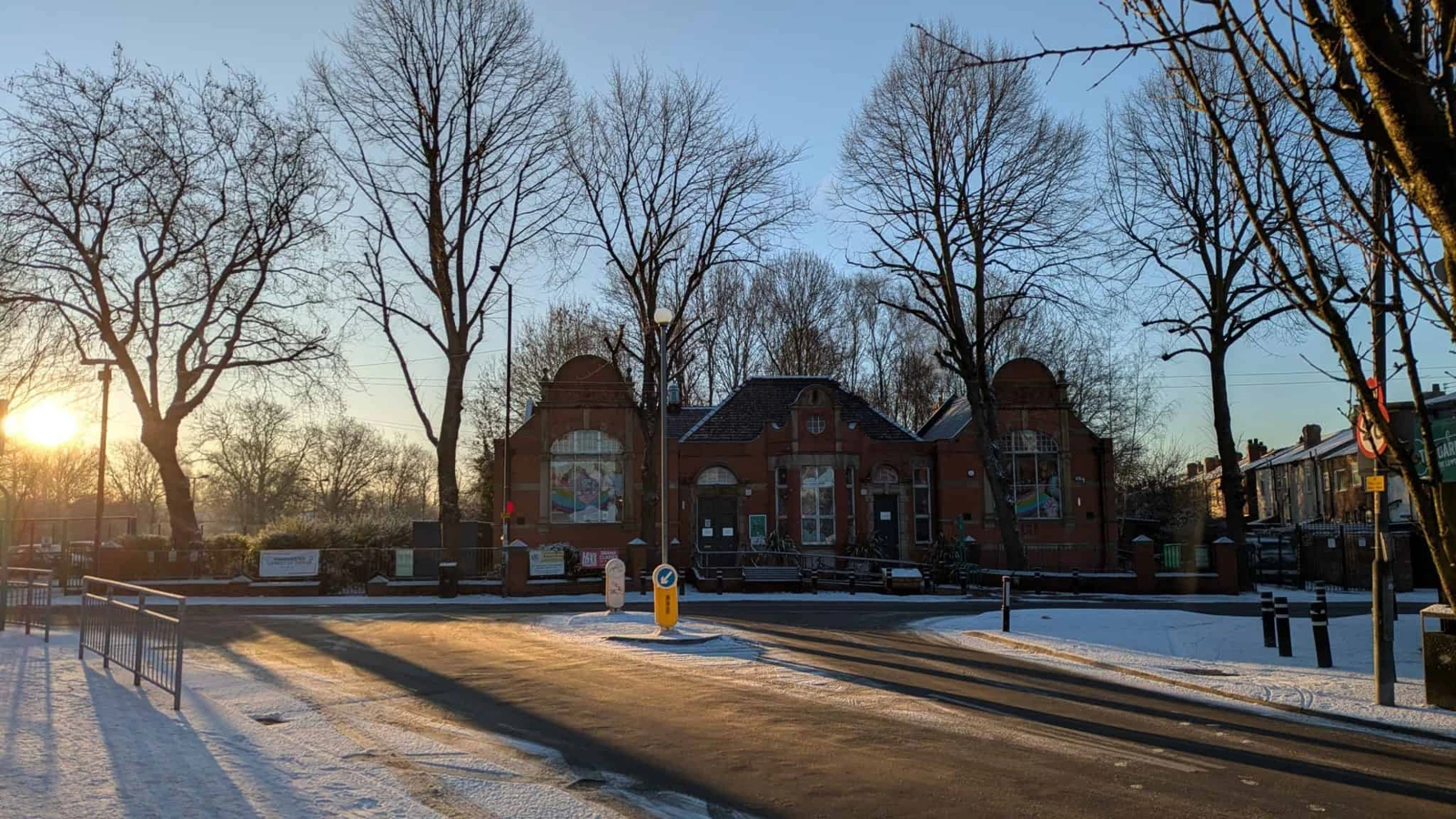 The red-brick Levenshulme Old Library building behind bare trees and a snow-dusted road in low winter sunlight