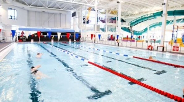 Lane-marked main swimming pool beneath a white roof at Andersonstown Leisure Centre, with slides behind
