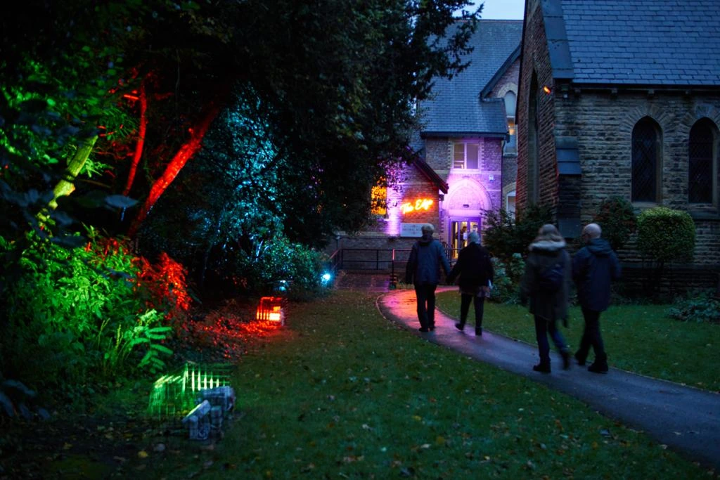 Visitors walking towards The Edge theatre entrance beside trees lit in coloured lights at dusk