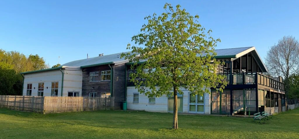 Bar Hill Village Hall beside a grassy field, with a leafy tree in front of its timber and pale-panelled exterior