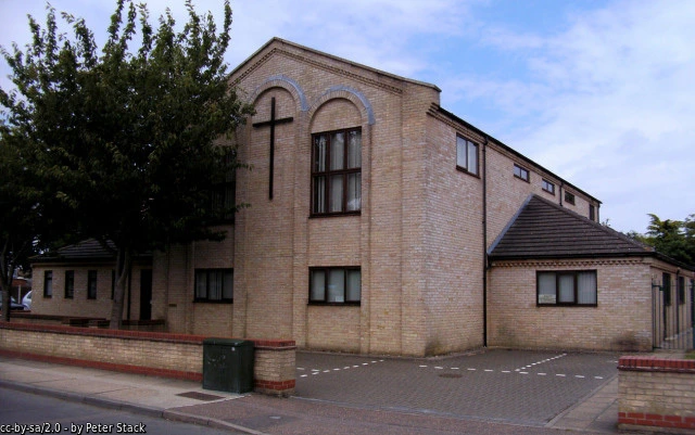 Brick exterior of Plume Avenue Church, with a cross on the front wall and trees beside the entrance