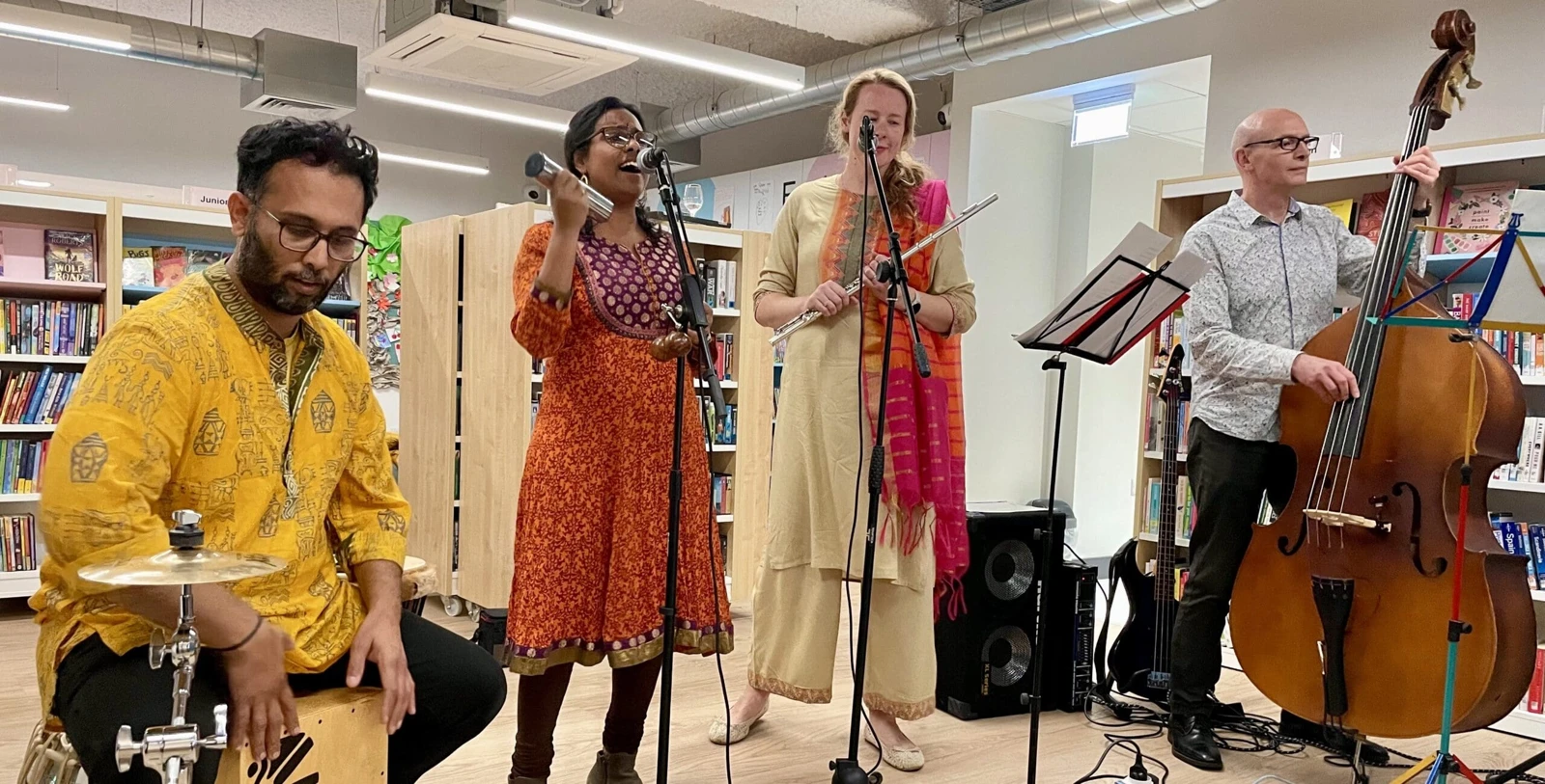 Four musicians performing with percussion, flute and double bass among library bookshelves at Cherry Hinton Hub
