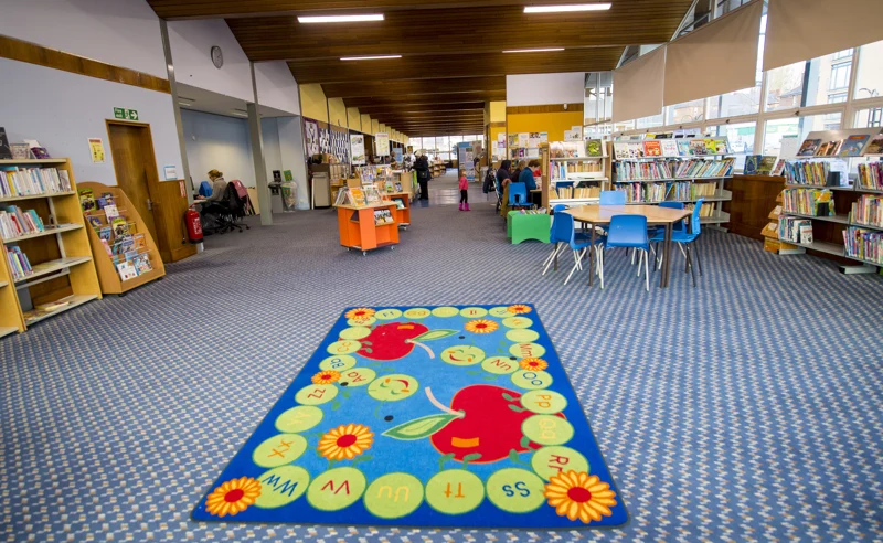 A colourful alphabet rug, blue chairs and book displays in the children’s area of Pollokshaws Library